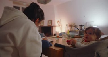 Anne yemek masasına eğilip oğluna okul ödevlerini yaptırırken küçük kardeş de elinde kırmızı oyuncakla evde oturmayı sembolize ediyor.