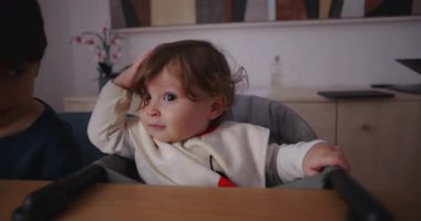 Aile yemeği sırasında yüksek sandalyede oturan gülümseyen çocuk arka planda kardeşlik yaparken neşeyle başını kaldırıyor ve evde özgün bir günlük dinamik ve aile bağı oluşturuyor..
