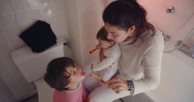 Anne, küçük banyoda çocukların dişlerini fırçalamasına yardım ediyor, büyük oğluna diş fırçasıyla yardım ederken, aile bakımında, günlük hijyende ve besleyici sabah rutininde.