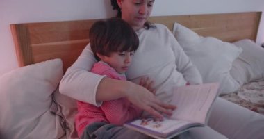 Anne yatakta oğlana masal kitabı okuyor. Sevecen bir uyku zamanı. Sessiz bir evde anne-baba ve çocuk arasında odaklanma, şefkat ve bağ gösteriyor.