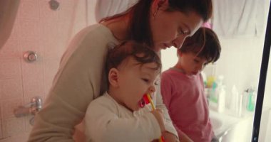 Anne küçük banyoda çocukların dişlerini fırçalamasına yardım ediyor, bebek diş fırçası çiğnerken büyük oğlu taklit ediyor, rutin, sıcak ve sevgi dolu ebeveynlik anları sergiliyor.