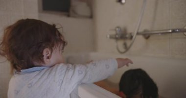 Küçük çocuk banyo sırasında küvette ağabeyine doğru uzanıyor. Kardeş etkileşimi, aile bağları ve ev banyosunda günlük rutinini gösteriyor..