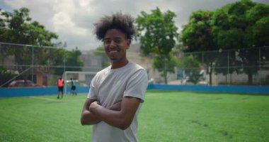 Futbol sahasında kendine güvenen, kollarını kavuşturmuş, takım arkadaşları oynarken gururunu, güvenini ve neşesini ifade eden Afro-Amerikalı Latin bir adam.