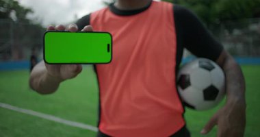 Afrika kökenli Latin Latin kökenli, elinde akıllı telefon, futbol sahasında yeşil ekran görüntüsü, uygulama veya sosyal medya teknolojisi ve modern yaşam tarzı olan Latin kökenli adamın yatay yakın çekimi.