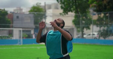 Afrika kökenli Amerikalı bir adam antrenman sırasında futbol sahasında kafa sallıyor, topu titizlikle kontrol ediyor ve şehir dışındaki çevreye odaklanıyor.