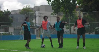 Bir grup erkek ısınma sırasında futbol sahasında esniyor ve tekmeliyor. Açık hava spor sahasındaki maçtan önce enerji, koordinasyon ve coşku gösteriyorlar.