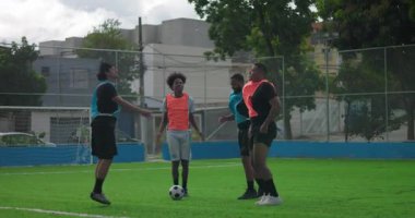 Futbol sahasında dört genç adam maç için hazırlanıyor, ısınıyor ve topun yanında esniyorlar, takım çalışması, dostluk gösteriyorlar ve arkadaşça bir maç yapmadan önce odaklanıyorlar.