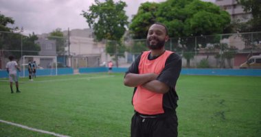 Futbol sahasında kolları kavuşturulmuş, turuncu yelek giyen, gururunu, yoldaşlığını ve motivasyonunu spor aracılığıyla ifade eden Afrikalı Amerikalı Latin bir adam.