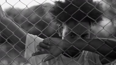 Latin kökenli Afrikalı bir adam siyah beyaz çitlere yaslanmış, hayal kırıklığı, acı ve duygusal yorgunluk anında yüzünü kollarıyla kapatıyor.