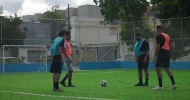 Yeşil sahada futbol oynayan bir grup erkek, ısınma antrenmanı sırasında takım arkadaşları arasında top oynuyorlar, şehir merkezinde takım çalışması, koordinasyon ve günlük arkadaşlık gösteriyorlar.