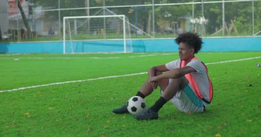 Afrika kökenli Latin kökenli genç bir adam çim futbol sahasında antrenmandan sonra futbol topuyla oturuyor. Şehir spor parkında gol ağlarına yakın düşünceli bir ifade.