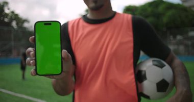 Latin kökenli, Afrika kökenli, elinde akıllı telefonuyla futbol sahasında top, teknoloji ve maket taşırken yeşil ekran görüntüsü olan Latin asıllı adamın yakın çekimi.