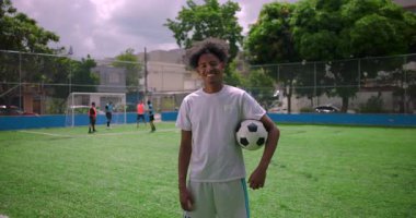Afrika kökenli Amerikalı Latin adam sahada futbol topunu tutarken güvenle gülümsüyor, takım arkadaşları antrenman yapıyor, takım çalışması kavramı, gurur, motivasyon ve toplum sporu ruhu.