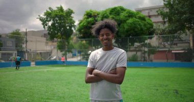 Futbol sahasında gururla gülümseyen Afrikalı Latin bir adam, kollarını kavuşturmuş rahat poz veriyor, arka planda takım arkadaşları, arkadaşlığın, takım çalışmasının ve sporda pozitif enerjinin sembolü.