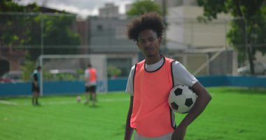 Afrika kökenli Brezilyalı bir adam futbol topunu tutarken ciddi görünüyor ve takım arkadaşları onun arkasında antrenman yaparken odaklanıyor.
