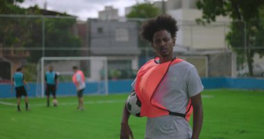 Afrika kökenli Latin kökenli bir adam futbol topunu sahada tutarken kameralar arka plandaki oyunculardan ona odaklanıyor. Kendine güvenen ifade ve güçlü varlık.