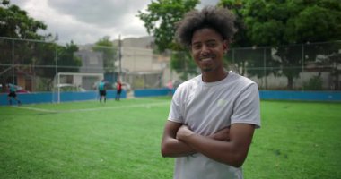 Futbol sahasında güvenle gülümseyen Afrikalı Latin bir adam, takım arkadaşları arkada oynarken kollarını kavuşturan, dostluk, gurur ve topluluk sporlarında neşe kavramı