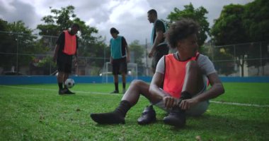 Arkadaşlarıyla gündelik futbol maçından önce bağcıklarını bağlayan genç Afrikalı Amerikalı adam, güneşli açık hava sahası, takım çalışması, odaklanma, beklenti ve spor rutinine hazırlanıyor.