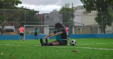 İspanyol bir adam futbol maçından önce çim sahasında oturuyor. Takım arkadaşı, ısınma sırasında takım çalışması, hazırlık ve karşılıklı destek göstererek ayağa kalkmasına yardım ediyor.