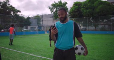 Futbol sahasında duran İspanyol adam elinde top tutarken akıllı telefonu kontrol ediyor. Spor antrenmanı sırasında teknolojiyle odaklanma ve günlük etkileşim gösteriyor.