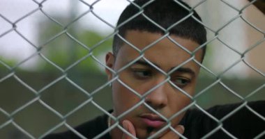 Eli çenesinde metal çitlerin arkasında dikilen Latin kökenli Latin kökenli bir adam. Dalgın bir yansımada kaybolmuş. Yalnızlığı, düşünceli ve sakin bir duygusal derinliği ifade ediyor.