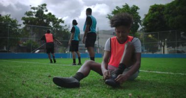 Arkadaşlarıyla gündelik futbol maçından önce bağcıklarını bağlayan genç Afrikalı Amerikalı bir adam sahada onlara katılmak için ayağa kalkıyor.
