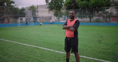 Futbol sahasında kollarını kavuşturarak sırıtan Afrikalı Latin bir adam, takım arkadaşları arkada oynuyor.