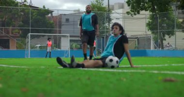 Takım arkadaşı antrenmandan sonra İspanyol futbolcuların ayağa kalkmasına yardım ediyor, dışarıda sahada antrenman yaparken oyuncular arasındaki cesaret, takım çalışması ve dayanışmayı temsil ediyor.