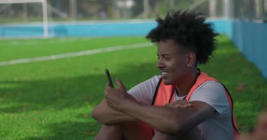 Afrikalı Amerikalı futbolcu, arkadaşlarıyla kamerasız sohbet ederken çimlerin üzerinde oturur, rahatlar ve kentsel alanda dostça sohbet ederken gülümserler.