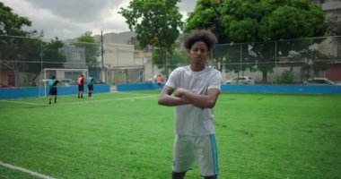 Futbol sahasında kolları çapraz duran Afrikalı Amerikalı Latin bir adam, ciddi ve odaklanmış bir ifade, arka planda oynayan takım arkadaşları