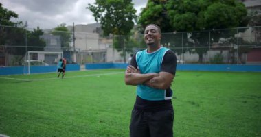 Latin kökenli, Afrika kökenli, futbol sahasında güvenle gülümseyen, takım arkadaşları arkada oynarken kollarını kavuşturan, arkadaşlığın, gururun ve halk sporlarında birliğin sembolü olan Latin bir adam.