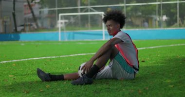 Afrika kökenli Brezilyalı bir adam futbol sahasında oturuyor, bacak bacak üstüne top oynuyor, düşünceli ve biraz hayal kırıklığına uğramış bir ifadeyle, gol ağının yakınına bakıyor.