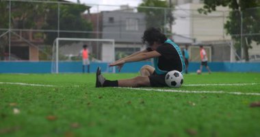 İspanyol bir adam antrenmandan önce futbol sahasında esniyor, topun yanına uzanmak için öne uzanıyor, takım arkadaşı ona yardım eli uzatıyor, motivasyon ve takım çalışmasının sembolü.