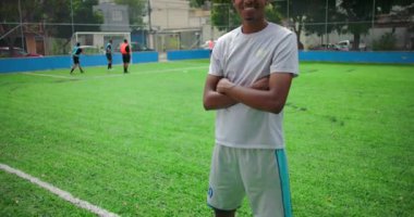 Afro-Amerikalı Latin adam futbol sahasında güvenle gülümsüyor, kollarını kavuşturmuş, takım arkadaşları arkada oynuyor, birlik, motivasyon ve takım çalışması kavramı