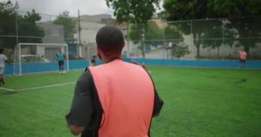 Afrika kökenli Latin erkek futbol sahasına arkadan giriyor, turuncu yelek giyiyor, takım arkadaşları hazırlanırken topa doğru koşuyor, hareket konsepti, takım çalışması ve motivasyon.