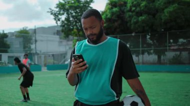 Futbol sahasında Afrikalı Amerikalı bir adam telefonu kontrol ediyor ve kameraya ekranı gösterirken gülümsüyor, açık havada rahatlamış bir bağlantı ve neşe anı yaşıyor.