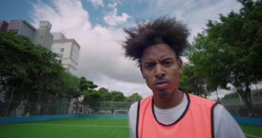 Turuncu önlüklü afro saçlı genç adam futbol sahasında duruyor, ciddi ifade, kararlılığı yansıtıyor ve şehir sporlarına odaklanıyor.