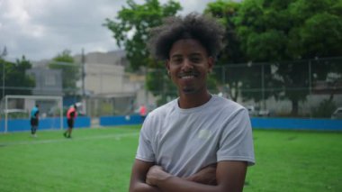 Açık hava futbol sahasında güvenle duran genç adam, kollarını kavuşturup gülümsüyor, atletizmi, kendine güveni ve spor ve takım çalışmasının keyfini temsil ediyor.