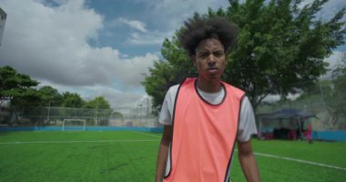 Futbol sahasında turuncu eğitim önlüğü giyen genç adam, ciddi ve kararlı ifade, afro saç stili, şehir spor ortamında odaklanma ve kararlılık gösteriyor