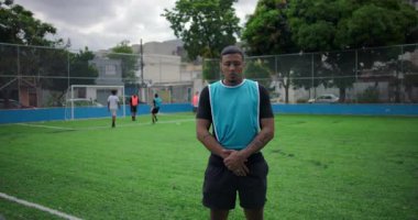 Arkadaşları arka planda, halk sporları ve takım ruhu konseptinde oynarken, genç Latin adam, dostluk maçı sırasında futbol sahasında güvenle duruyor, kameraya bakıyor.