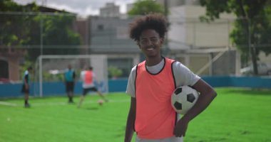 Afrika kökenli Brezilyalı bir adam futbol sahasında, takım arkadaşları antrenmandan sonra neşe ve takım ruhunu ifade ederken, top koltukaltında güvenle gülümsüyor.