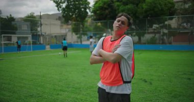 Latin asıllı bir adam futbol sahasında kollarını kavuşturmuş, ciddi ve odaklanmış görünüyor, açık hava futbol antrenmanı sırasında kararlılık, güven ve hazırlık gösteriyor.