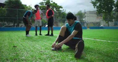Gündelik futbol maçından önce arkadaşlarıyla ayakkabı bağı bağlayan genç bir İspanyol, güneşli açık hava sahası, takım çalışması, odaklanma, beklenti ve spor rutinine hazırlanıyor.