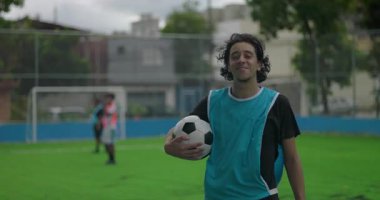 İspanyol adam takım antrenmanı sırasında sahada güvenle gülümseyen futbol topunu tutuyor, takım arkadaşları şehir parkında antrenman yaparken pozitifliğini ve gururunu dile getiriyor.