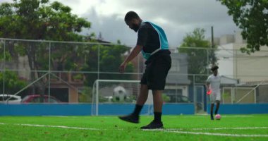 Kara bulutlu gökyüzü altında antrenman öncesi futbol sahasında ısınan Afrikalı Amerikalı bir adam, şehir parkındaki egzersiz sırasında denge ve top kontrolüne odaklanıyor.