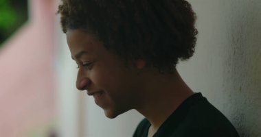 Olumlu bir ifadeyle gülümseyen, duvara dayanan, Afrika kökenli, açık hava manzaralı, mutluluk ve memnuniyet dolu, yakın çekim profilli bir çocuk.