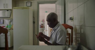 Bir yaşlı siyah adam, sade bir mutfakta yemek masasında oturuyor, akıllı telefon kullanıyor, günlük hayatta sıcak ve tanıdık bir ortamda teknolojiyi ön plana çıkarıyor.
