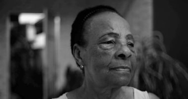 Düşünceli bir profilde yaşlı bir Afro kökenli kadın, kameradan başka tarafa bakıyor, açık bir ev ortamında düşünceli bir yüz ifadesi, doğal ve samimi.