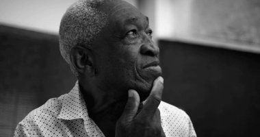 Siyah beyaz bir portre içinde yaşlı bir Afro-torun adam, düşünceli bir gülümseme, el dokunuşlu çene ile yukarı bakıyor, yüz hatları açık bir evde vurgulanmış