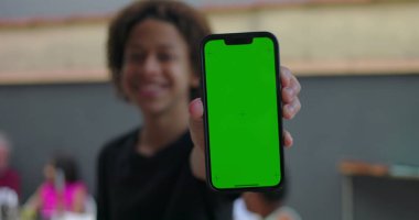Afrika kökenli bir gencin elinde yeşil ekranlı akıllı telefon, krom anahtar ekranlı, özgüvenle gülümseyen, arka planda çeşitli yetişkinlerle gelişigüzel toplanan